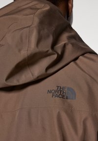 Barna vízálló dzseki kapucnival, texturált felülettel és a "The North Face" fekete logóval a jobb vállon.