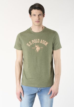 Uomo in piedi che guarda verso il fronte, indossa una t-shirt verde oliva U.S. Polo Assn. e jeans azzurri chiari su sfondo bianco.