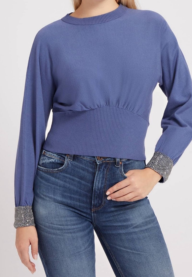 Woman wearing a purple long-sleeve knit top with rhinestone cuffs tucked into high-waisted blue jeans, hand resting on hip.