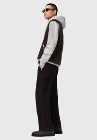 Gilet di pile marrone sopra un hoodie grigio, abbinato a pantaloni cargo neri larghi e scarpe nere. Gli accessori includono occhiali da sole rettangolari.
