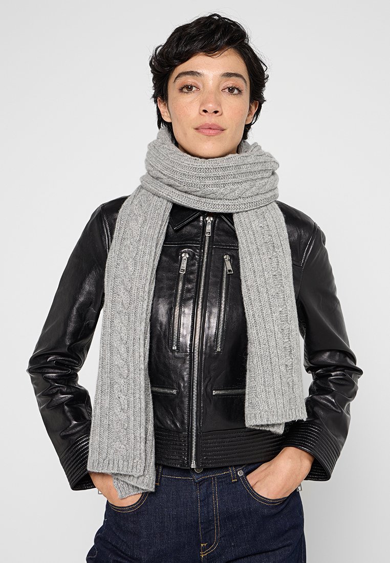 Szary dziany szalik z wzorem w warkocze, noszony luźno na szyi na czarnej skórzanej kurtce. Szalik jest miękki i ma teksturę.