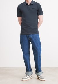 Ciemnoszara koszulka polo z dwoma guzikami, w połączeniu z podwiniętymi niebieskimi dżinsami i szarymi trampkami z białymi akcentami i paskowanym wzorem.