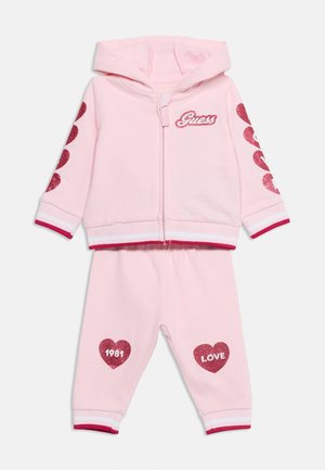 Ensemble rose pour tout-petit avec sweat à capuche et pantalon zippés, cœurs rouges pailletés sur les manches et les genoux, logo "Guess" sur la poitrine et textes "1981" et "LOVE".