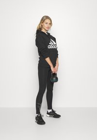 Haut à capuche noir avec le logo Adidas blanc, leggings noirs avec des rayures blanches, et chaussures de sport noires. Tenant un kettlebell noir.