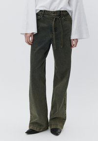 Olivengrønne jeans med vide ben lavet af denim, med høj talje, forlommer og en snøre i taljen. Sorte sko med spids tå.