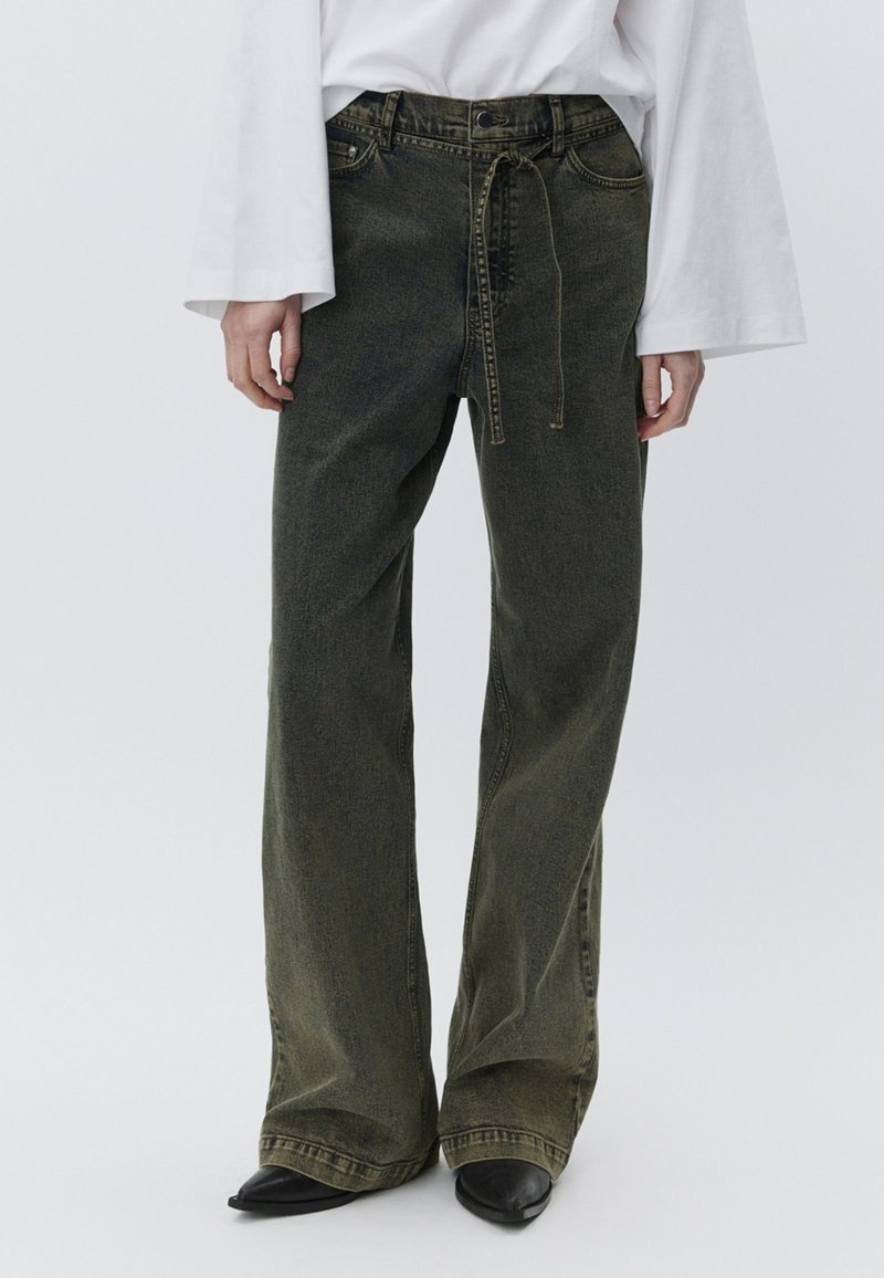 Olivengrønne jeans med vide ben lavet af denim, med høj talje, forlommer og en snøre i taljen. Sorte sko med spids tå.