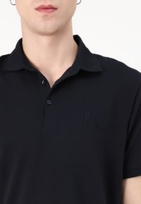 Námornícka polo košeľa s krátkymi rukávmi, so zapínaním na gombíky a textúrovaným vyšitým logom "K" na hrudi. Mäkká tkanina s hladkým povrchom.