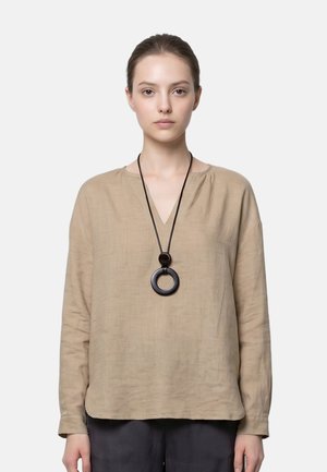 Giovane donna che indossa una camicetta beige a maniche lunghe e una grande collana nera con ciondolo circolare contro uno sfondo chiaro e uniforme.