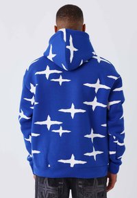 Sudadera azul con un patrón estelar abstracto blanco, con capucha ajustable y puños acanalados. Textura de tela suave y corte holgado.