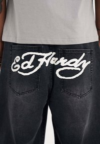 Mustat farkut, joissa on suuri valkoinen "Ed Hardy" -logo ommeltuna takataskujen yli, perinteiset taskusuunnittelut ja ompeluyksityiskohdat.