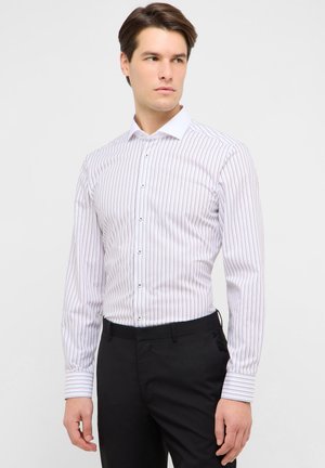 Mand iført hvid langærmet skjorte med sorte lodrette striber og sorte bukser, der kigger mod venstre på en ensfarvet baggrund.