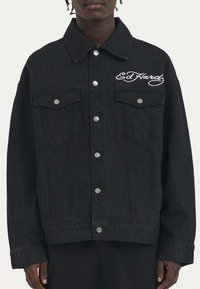 Giacca in denim nera con una vestibilità leggermente over size, caratterizzata da bottoni a pressione argentati, tasche sul petto e logo del marchio ricamato sul lato sinistro.