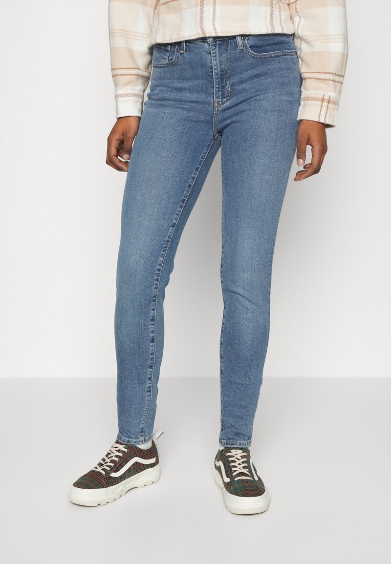 Personne portant un jean skinny bleu clair, une chemise à carreaux beige et des baskets marron à motifs, debout devant un fond clair uni.