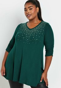 Vihreä tunika, jossa on V-pääntie ja kolme neljäsosaa hihat. Vahvistettu hopeisilla helmin koristeluilla kaula-aukossa ja rento, virtaava istuvuus.