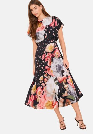 Femeie purtând o rochie midi neagră cu imprimeu floral mare multicolor, mâneci scurte și sandale negre cu toc înalt și barete, stând în picioare pe un fundal alb.