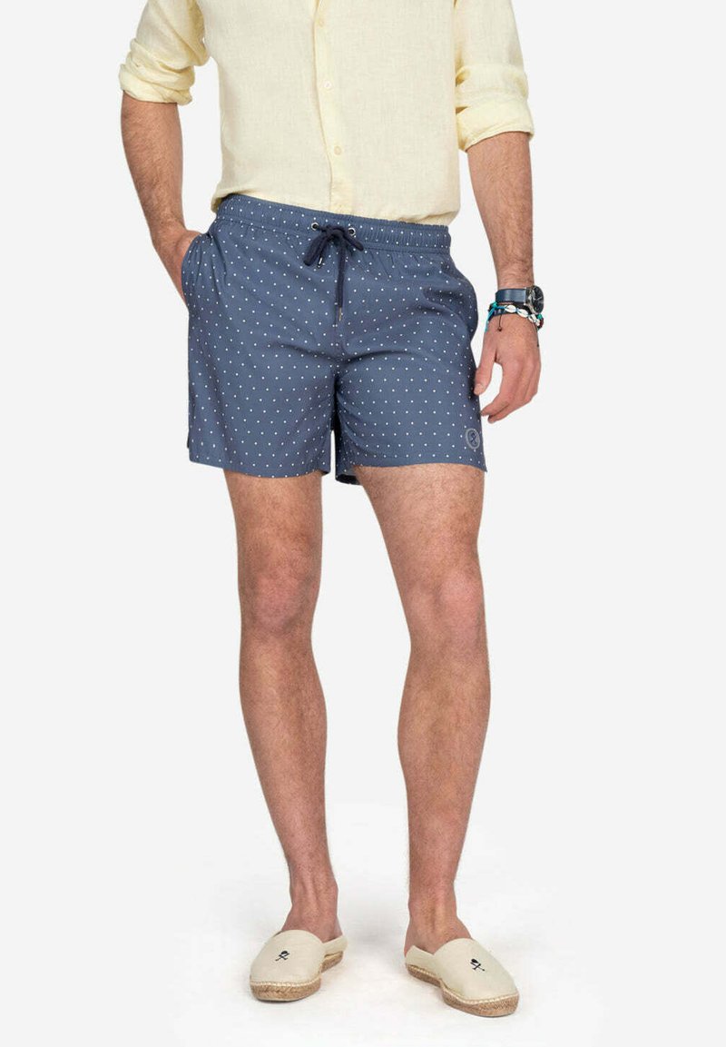 Bañador azul con lunares, con cintura elástica y cordón de ajuste, hecho de tejido ligero. Llevado con alpargatas beige y una camisa ligera.