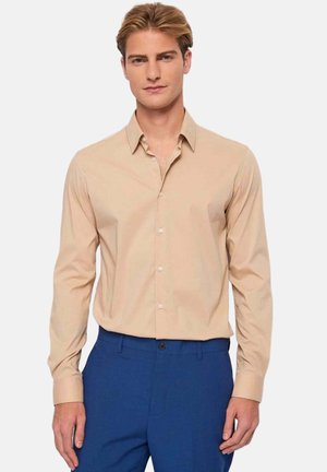 Giovane uomo che indossa una camicia beige con bottoni infilata in pantaloni eleganti blu, in piedi contro uno sfondo chiaro e semplice.