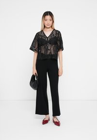 Top de encaje negro con mangas cortas y un patrón ondulado, combinado con pantalones rectos negros y zapatos planos rojos. La modelo sostiene un bolso negro.
