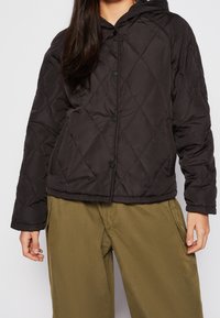 Schwarze gesteppt Jacke mit Kapuze, ausgestattet mit Druckknopfverschluss und gerippten Bündchen, kombiniert mit lockeren olivgrünen Hosen.