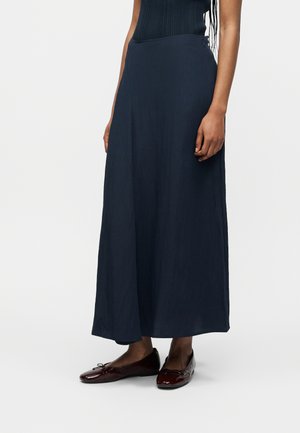 Femme portant une longue jupe bleu marine foncé associée à des ballerines rouge foncé, debout devant un fond blanc uni.