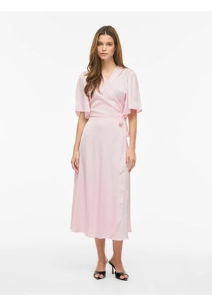 Kobieta w jasnoróżowej sukience kopertowej z falbanowymi rękawami i czarnych sandałkach na obcasie, stojąca na białym tle.