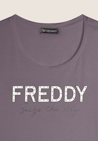 T-shirt violet à manches courtes avec encolure dégagée, portant des lettres majuscules cloutées blanches avec le mot "FREDDY" et la phrase "Saisis le jour" en dessous.