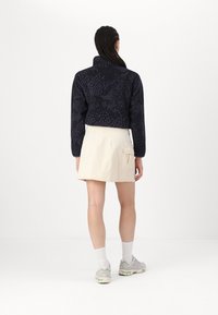 Kortad marinblå fleecejacka med texturerat mönster, hög krage; bärs med en beige kjol med sidoficka. Ljust grå sneakers.