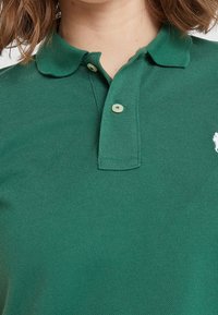 Grön polo skjorta gjord av texturerad tyg, med en klassisk krage, två knappar och en vit logotyp på vänster bröstområde.