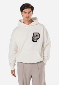 Hoodie de cor creme com um bolso frontal, apresentando um emblema texturizado em preto e prateado. Ajuste suave e relaxado com capuz ajustável.