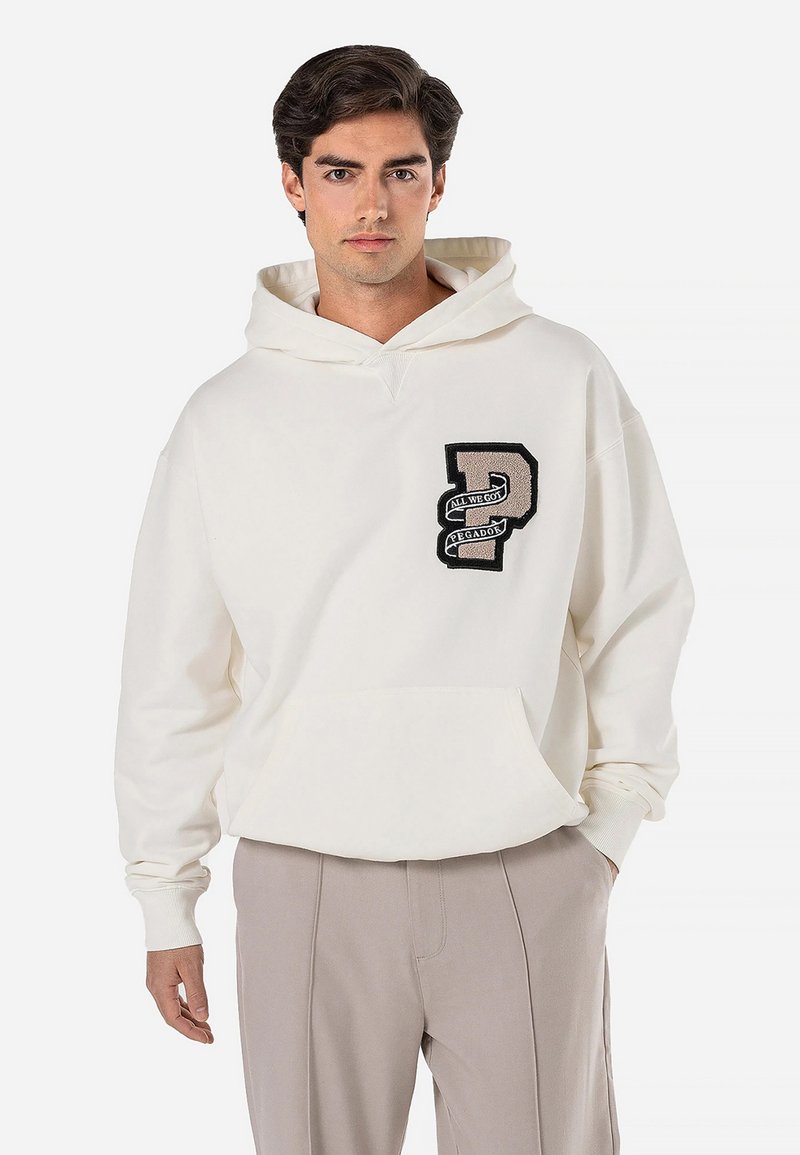 Hoodie de cor creme com um bolso frontal, apresentando um emblema texturizado em preto e prateado. Ajuste suave e relaxado com capuz ajustável.