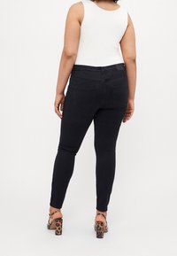 Černé džíny skinny s středně vysokým pasem, zadními kapsami a úzkým střihem, spojené s bílým topem bez rukávů a vzorovanými podpatky.