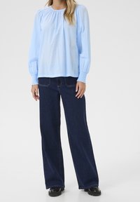 Vaaleansininen, pitkähihainen pusero, jossa on pistekuvioitu tekstuuri ja smokkaukset hihansuissa, yhdistetty tummansinisiin leveälahkeisiin farkkuihin ja mustiin kenkiin.