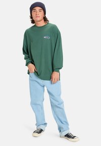 Jonge man met een marineblauwe muts, een donkergroen shirt met lange mouwen en een klein logo, lichtblauwe jeans en zwarte sneakers, staand met één hand in zijn zak.