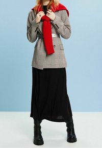Grau kariertes Blazer mit doppelter Knopfleiste, tailliert; kombiniert mit einem schwarzen Maxirock und einem roten Strickschal, ergänzt durch schwarze Stiefel.