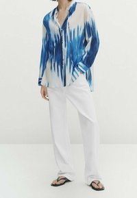 Przezroczysta koszula zapinana na guziki w kolorze niebieskim i białym z wzorem tie-dye, długimi rękawami i luźnym krojem, zestawiona z białymi spodniami o szerokich nogawkach i klapkami.