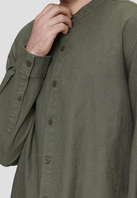 Man in olijfgroene, knoopjeshemd met lange mouwen, hand aan de kraag, voor een effen lichtgrijze achtergrond.