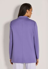 Vrouw met lang donker haar die wegkijkt, gekleed in een strakke lavendelkleurige blazer en witte broek tegen een effen lichte achtergrond.