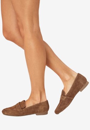 Jambes portant des mocassins en daim marron avec un petit talon et une sangle texturée sur fond blanc.