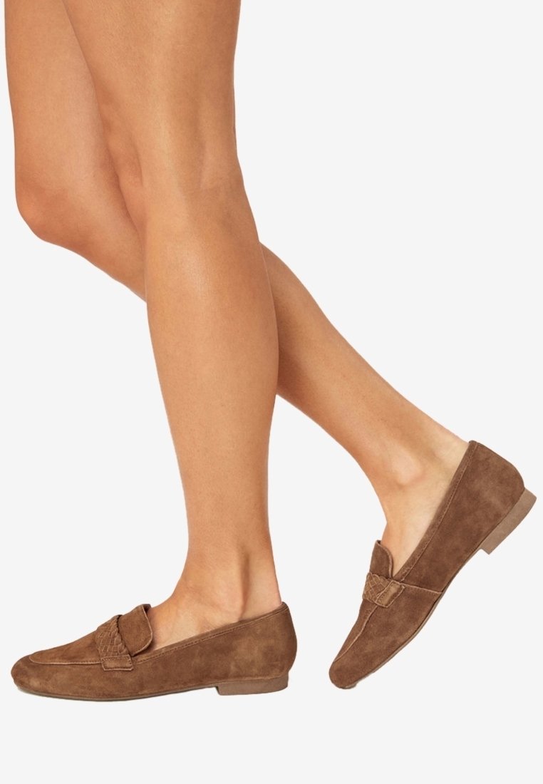 Jambes portant des mocassins en daim marron avec un petit talon et une sangle texturée sur fond blanc.