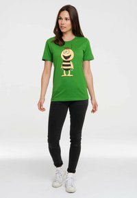 Green cotton t-shirt featuring a cartoon bee design with yellow body and black stripes. Paired with black jeans and white sneakers.