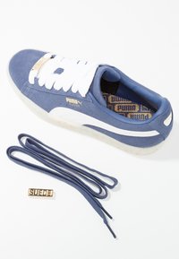 Baskets en daim bleu Puma avec une bande blanche et des lacets blancs, des lacets bleus supplémentaires, et une étiquette dorée "Suede" sur les lacets, sur fond blanc.