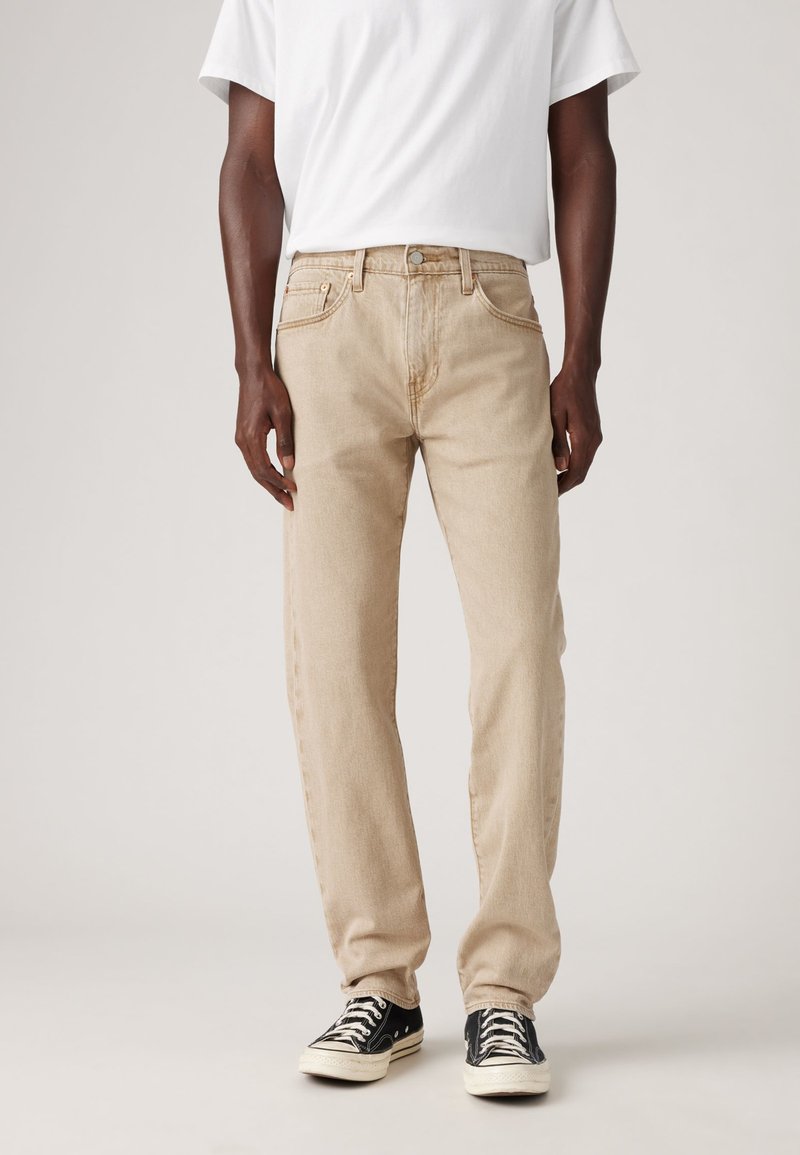 Beige denim jeans met een rechte pasvorm, voorzien van gestikte zakken en een knoopsluiting. G gedragen met zwarte sneakers en een wit t-shirt.