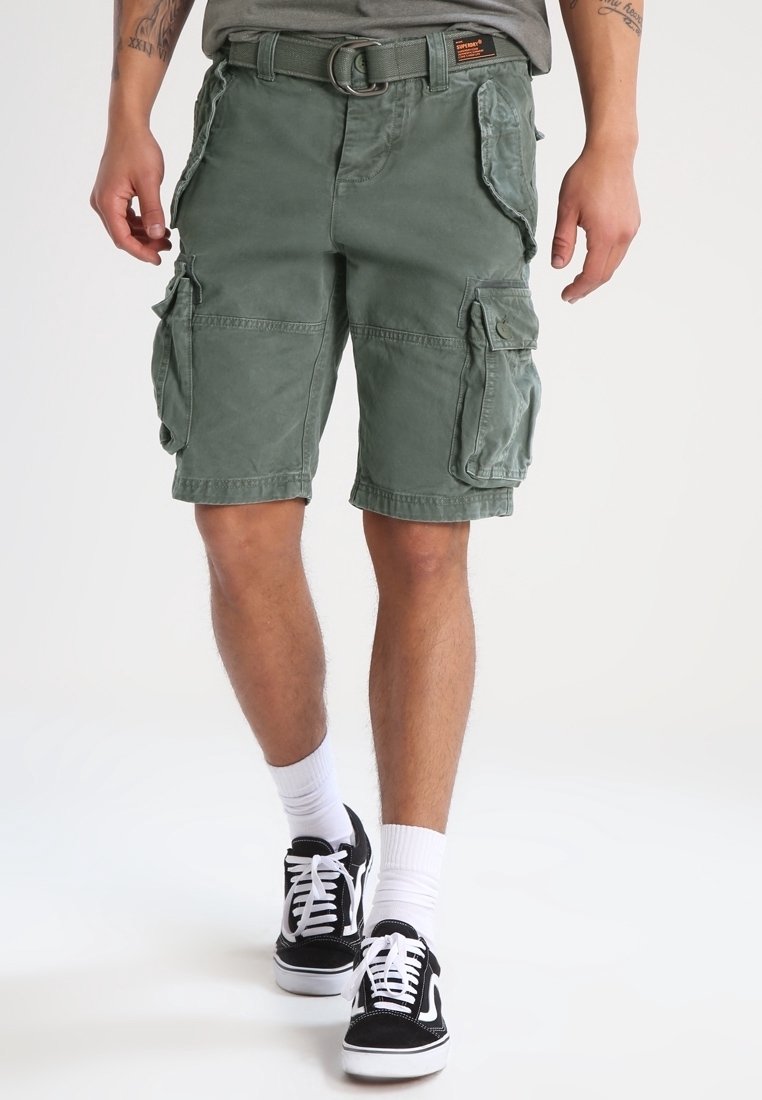 Homme portant un short cargo vert avec plusieurs poches, des chaussettes blanches et des baskets noires et blanches marchant vers l'avant sur un fond uni.