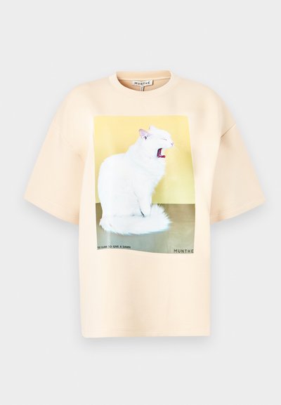 Oversized beige T-shirt van zachte stof met een grote afbeelding van een gapende witte kat op een gele en groene achtergrond, met tekst eronder.