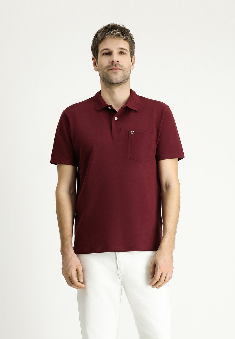 Polo di colore bordeaux con colletto, maniche corte e taschino sul petto. Caratterizzato da un piccolo dettaglio del logo. Abbinato a pantaloni bianchi.