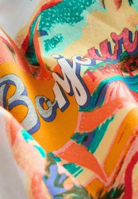 Bunt gemustertes Stoffdesign mit tropischem Motiv und fett gedrucktem Text, überwiegend in Orange, Grün und Pink, mit glatter Textur und lebhaften Mustern.
