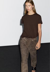 Brun bomulds t-shirt parret med taupe ruskindsbukser med synlig syning. Modellen bærer en sort sandal og en stor ring. Minimalistisk baggrund.