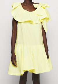 Robe courte jaune avec un col à volants et un grand nœud sur une épaule portée par une personne à la peau foncée sur un fond uni.