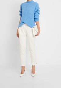 Blauwe gebreide trui met een hoge hals en geribbelde textuur, gecombineerd met crème tapered broek en witte puntige hakken. Klein wit clutch-tasje.