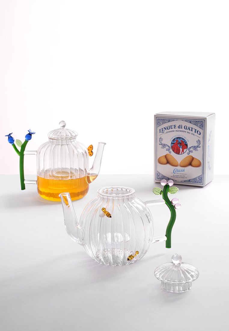 Heldere glazen theepotten met ribbelontwerp, kleurrijke bloemenaccenten en een doos koekjes. Eén pot bevat amberkleurige thee, die door de transparante zijkanten zichtbaar is.